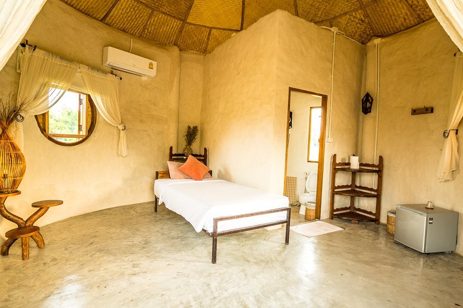 A small adobe bungalow at Mala Dhara — single bed, curved earthen walls and a round window onto the fields
